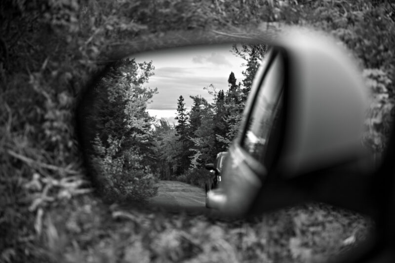 Schwarzweiß-Fotografie: Ein Blick in den Seitenspiegel des Autos zeigt was Hinter uns liegt, zeitgleich geht der Blick nach vorne. Dadurch, dass das Motiv im Spiegel heller ist, wird der Vordergrund dunkler abgelichtet.