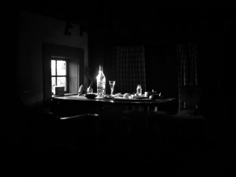 Schwarzweiß-Fotografie: Der Blick in ein Haus im Freilichtmuseum Skansen auf einen Tisch, der durch ein anderes Fenster und dessen einfallenden Lichtes beleuchtet wird. Da die Belichtung auf die helle Flasche und Gläser liegt, bildet der ansonsten dunkle (negative) Raum den Rahmen.